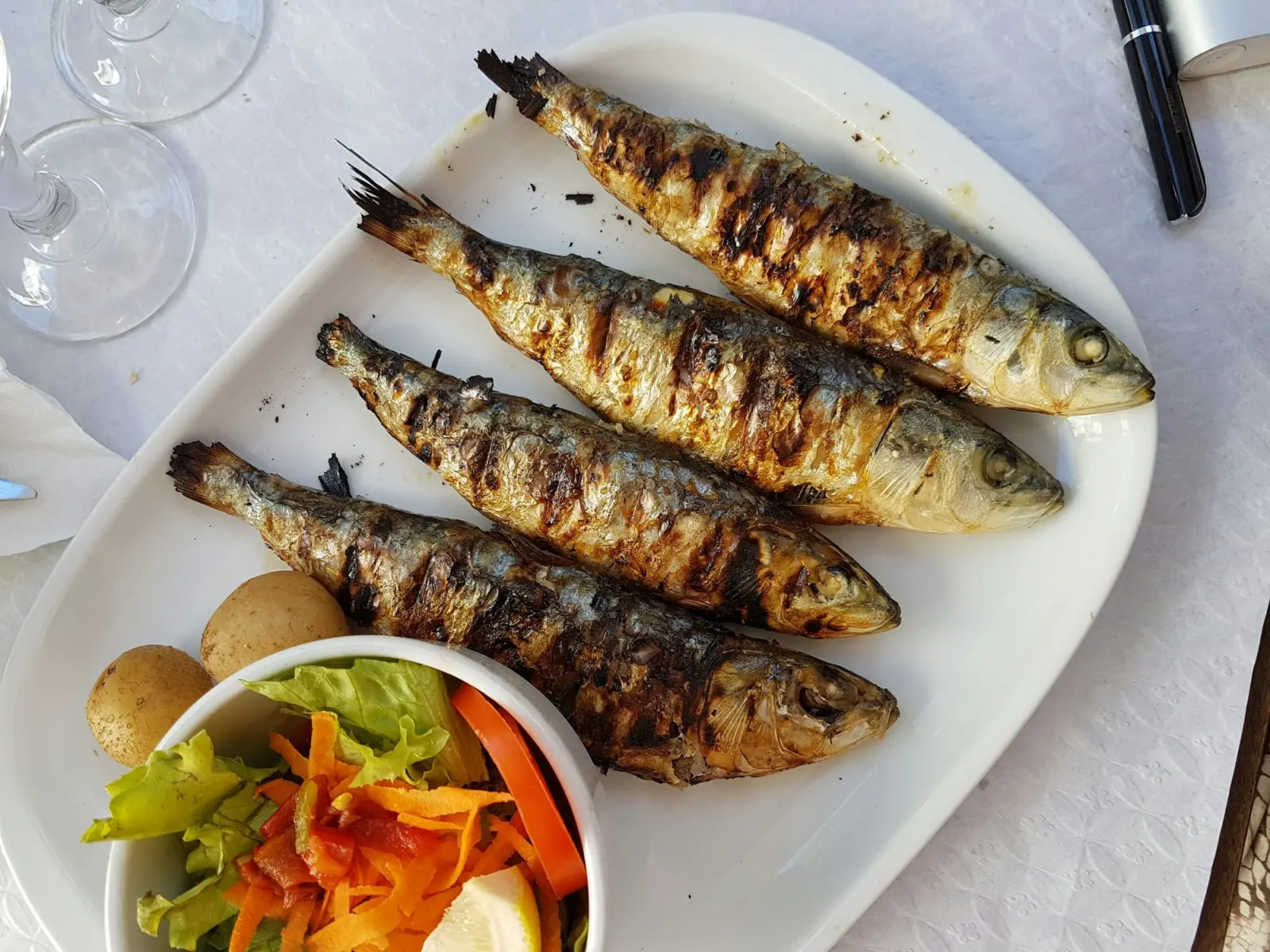 Travessa com quatro sardinhas assadas, batata cozida e salada de pimentos e cenoura.